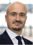 Mężczyzna w garniturze, białej koszuli i ciemnym krawacie, portret od ramion w górę, jasne rozmyte tło.