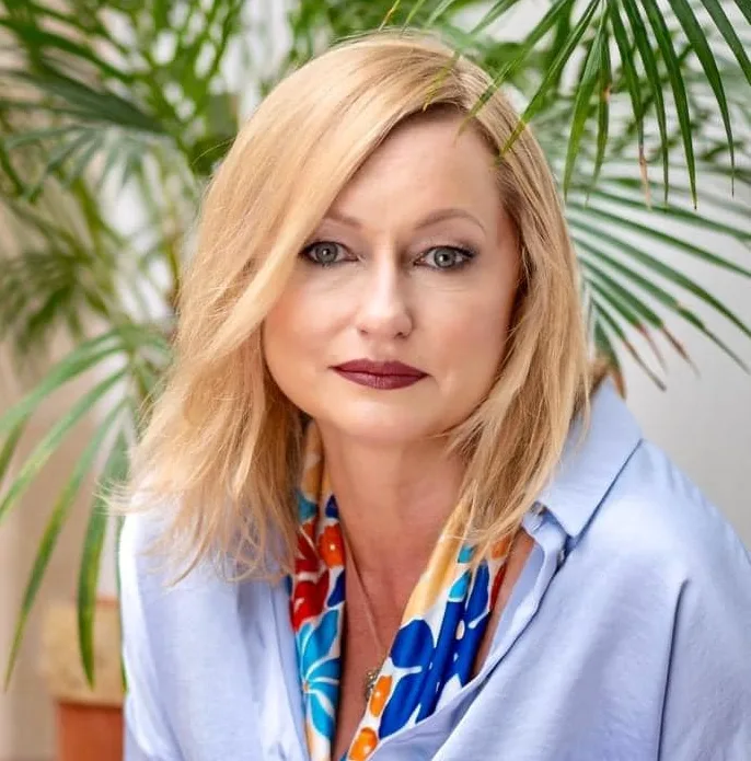 Kobieta w średnim wieku o blond włosach i jasnych oczach, ubrana w jasnoniebieską koszulę i kolorową apaszkę, siedząca na tle zielonych roślin.