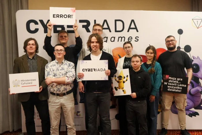 Grupa osób pozuje do pamiątkowej fotografii na tle ścianki z napisem CYBERIADA games. Uczestnicy trzymają w rękach tabliczki oraz materiały z logotypami i hasłami, w tym logo Ministerstwa Cyfryzacji, napis „CYBERIADA”, „ERROR!” oraz zabawne hasło „Moja gra działa... W teorii Lema wszystko działało!”.