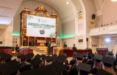 Rektor UEP przemawia z mownicy na scenie reprezentacyjnej sali z organami w tle, nad którą wisi duży baner z napisem Absolutorium / Graduation Ceremony. Na pierwszym planie widoczny jest widok z tyłu na zgromadzonych absolwentów w czarnych biretach.