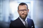 Portret biznesowy mężczyzny w garniturze, z brodą i w charakterystycznych okularach w grubych oprawkach, stojącego w jasnym wnętrzu biurowym.