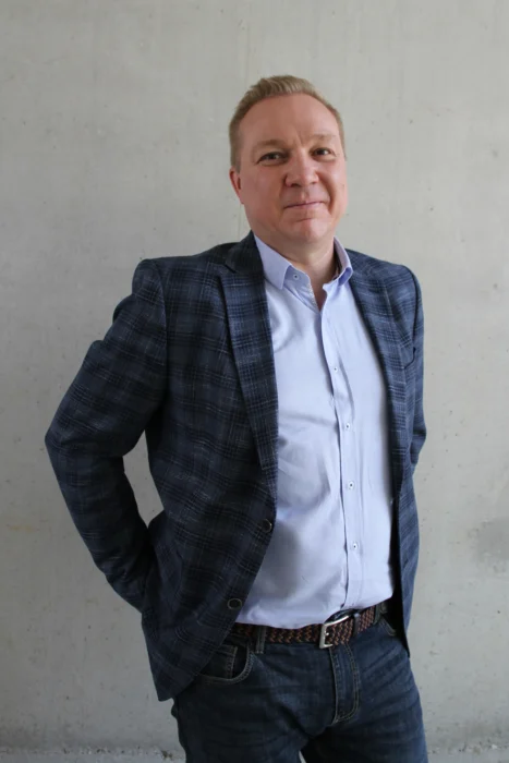 Mężczyzna o jasnych, krótkich włosach, ubrany w ciemnoniebieską marynarkę w kratę, błękitną koszulę oraz jeansy z plecionym paskiem, pozujący na tle szarej, betonowej ściany z rękami trzymanymi z tyłu.