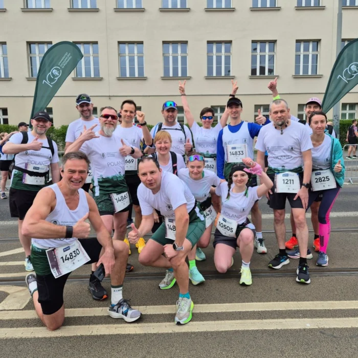 Grupa uśmiechniętych biegaczy w białych koszulkach z numerami startowymi i logo Uniwersytetu Ekonomicznego w Poznaniu, pozująca do pamiątkowego zdjęcia na ulicy na tle budynku uczelni.