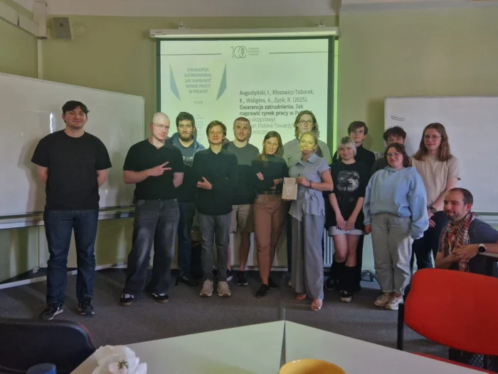 Grupa osób pozuje do pamiątkowej fotografii w sali wykładowej. W tle na ekranie wyświetlana jest prezentacja dotycząca książki „Gwarancja zatrudnienia. Jak naprawić rynek pracy w Polsce?”. Jedna z osób w pierwszym planie trzyma w ręku egzemplarz omówionej publikacji.